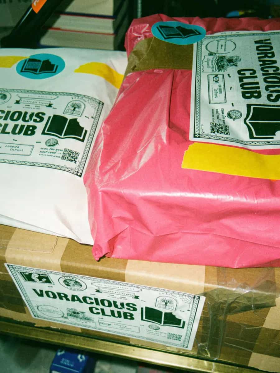 A white mailer and a pink parcel on a box, all with Voracious Club labels and stickers