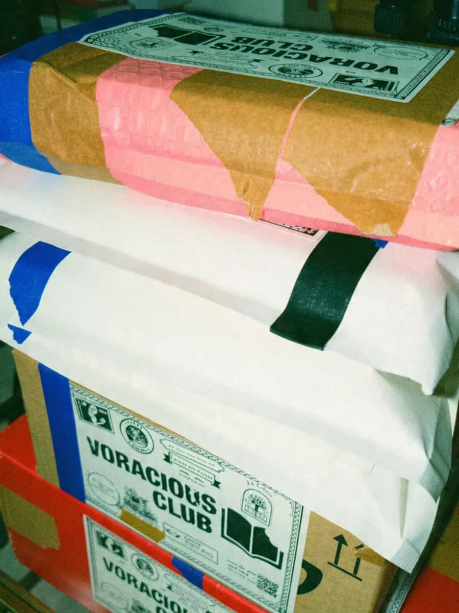 A tall stack of Voracious Club parcels: a pink bubble mailer, white envelopes and labelled boxes